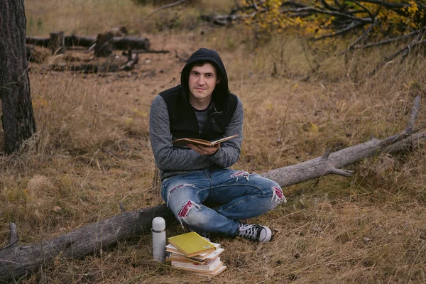Un joven guapo vestido de civil está sentado en un tronco y leyendo un libro en el bosque de otoño.. — Foto de Stock