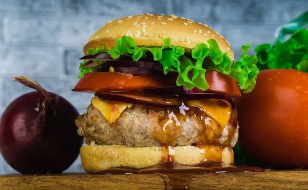 Um hambúrguer suculento enorme está em uma tábua de corte com legumes em um fundo de tijolo cinza . — Fotografia de Stock