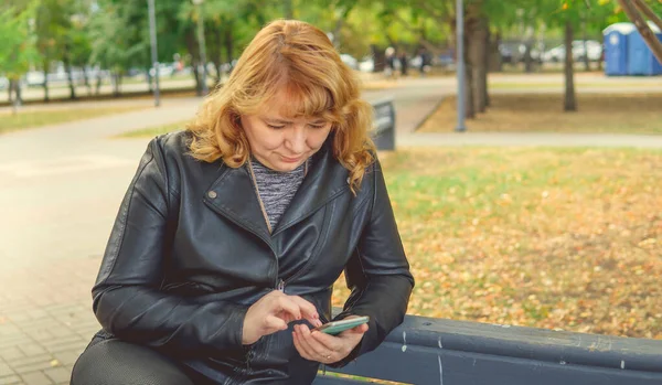 Een volwassen vrouw zit op de bank in de herfst Park draait het nummer op mobiele telefoon — Stockfoto