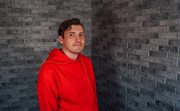 A young handsome man in a red hoodie with bristles poses on a gray brick background. — Stock Photo, Image