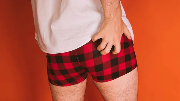 Male body in the plaid boxers and in white shirt on orange background. Mans hand scratches his ass. — 스톡 사진