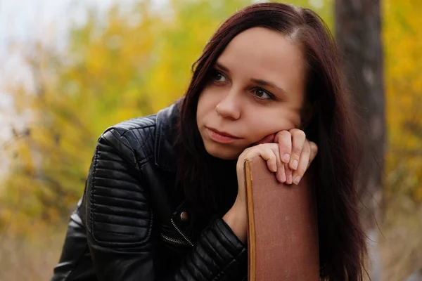 Uma jovem mulher bonita com cabelos escuros longos em roupas casuais está sentada em um log, segurando um livro em suas mãos, e cuidadosamente olhando para o lado. — Fotografia de Stock