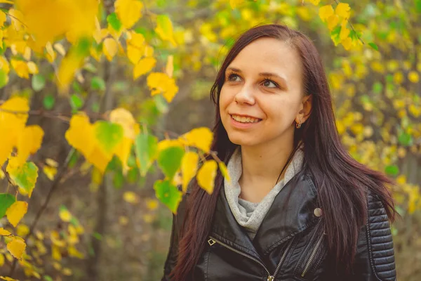 Retrato de uma mulher sorridente bonita está em pé na floresta de outono entre os ramos de árvores com folhagem amarela. — Fotografia de Stock