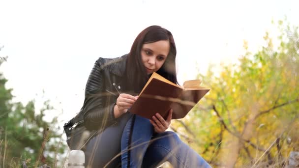 En ung vacker kvinna med långt mörkt hår i avslappnade kläder sitter på en stock och läser en bok i höstskogen. — Stockvideo