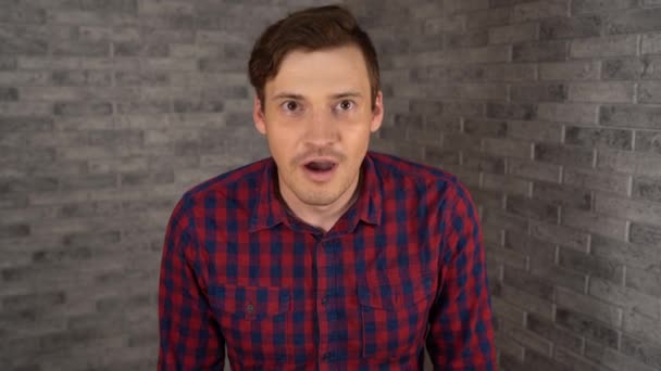 Um homem bonito novo na camisa xadrez abre sua boca e espalha suas mãos da surpresa em um fundo cinzento do tijolo. — Vídeo de Stock