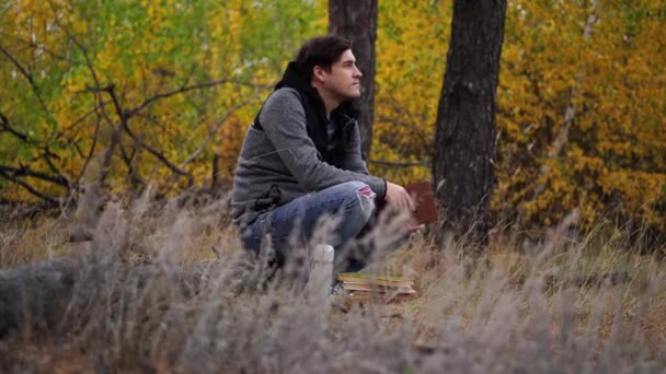 Ένας νεαρός όμορφος άντρας με καθημερινά ρούχα κάθεται σε ένα κούτσουρο και διαβάζει ένα βιβλίο στο δάσος του φθινοπώρου.. — Αρχείο Βίντεο