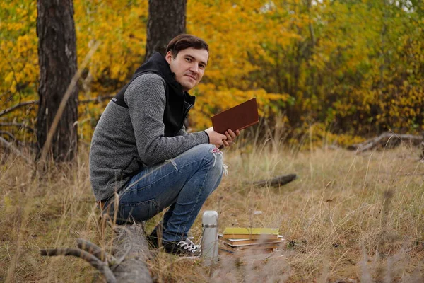 Mladý pohledný muž v neformálních šatech sedí na kládě a čte si knihu v podzimním lese. — Stock fotografie