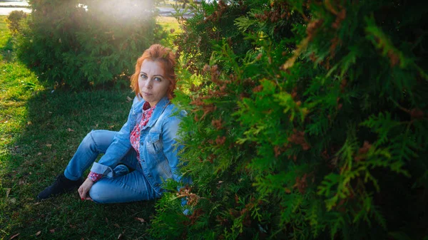 Jovem com cabelos vermelhos entre as árvores. Retrato de jovem mulher atraente com penteado moderno de pé no jardim da primavera e olhando para a câmera — Fotografia de Stock