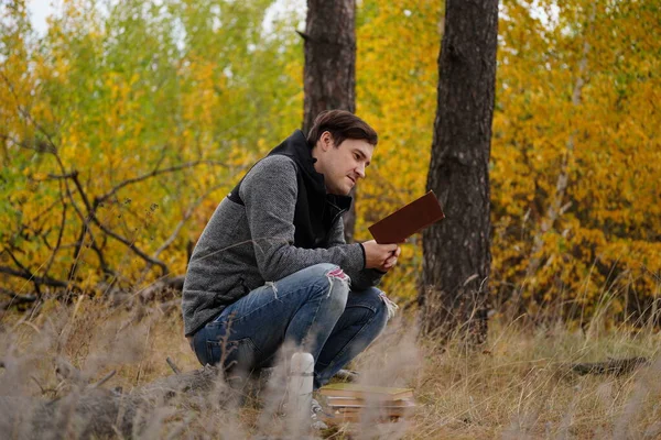 Un jeune et beau homme en tenue décontractée est assis sur un rondin et lit un livre dans la forêt d'automne.. — Photo