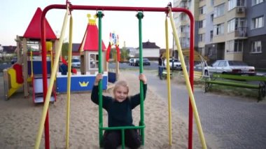Renkli çocuk parkında sallanan kız. Sıcak elbiseli genç kız salıncakta sallanıyor ve kameraya bakıyor.