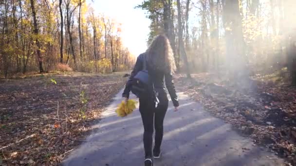 Névtelen nő sétál a parkban. Vissza kilátás fiatal nő díszítő hátizsák séta az úton a csendes őszi parkban — Stock videók