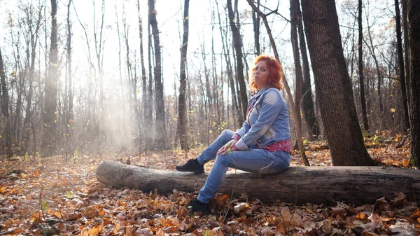 Mujer sentada en madera en el bosque de otoño. Mujer joven con pelo rojo vestida de civil sentada sobre troncos caídos en el bosque rural mirando hacia atrás bajo la luz del sol. — Foto de Stock