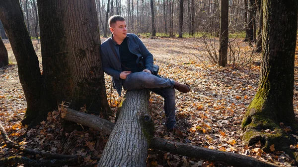 Hombre sentado en el tronco en los bosques de primavera. Hombre con ropa de vestir sentado en madera caída en el bosque rural mirando hacia atrás bajo la luz del sol. — Foto de Stock