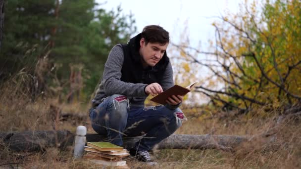 Un joven guapo vestido de civil está sentado en un tronco y leyendo un libro en el bosque de otoño.. — Vídeos de Stock