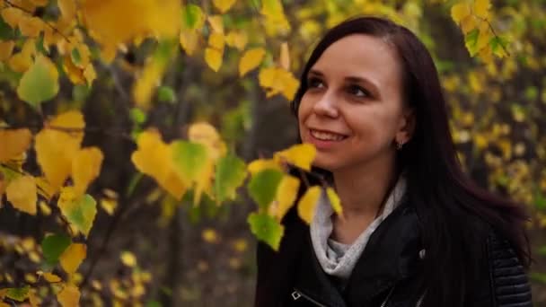 Портрет красивой улыбающейся женщины стоит в осеннем лесу между ветвями деревьев с желтой листвой. — стоковое видео