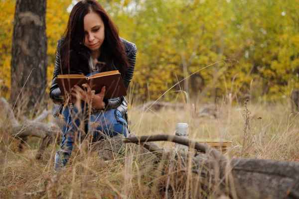 Egy fiatal, gyönyörű nő hosszú, sötét hajjal alkalmi ruhában ül egy fatörzsön és könyvet olvas az őszi erdőben.. — Stock Fotó