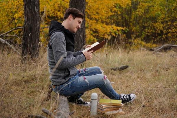 Egy fiatal jóképű férfi alkalmi ruhában ül egy fatörzsön és könyvet olvas az őszi erdőben.. — Stock Fotó