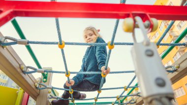 Sıradan giyinmiş sevimli küçük bir kız şebekede oturur ve şehir merkezindeki oyun parkındaki kameralara bakar..