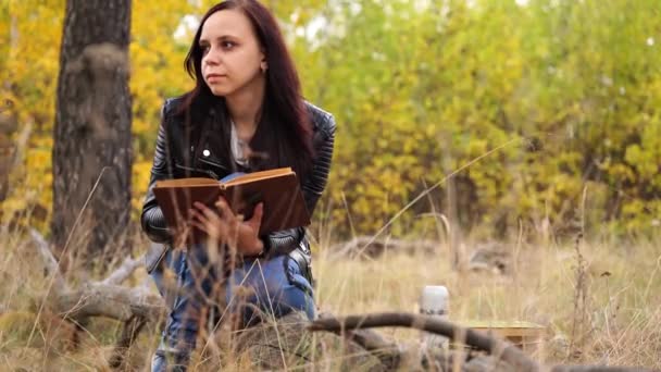 Une jeune et belle femme aux cheveux longs et foncés en tenue décontractée est assise sur un rondin et lit un livre dans la forêt d'automne. — Video