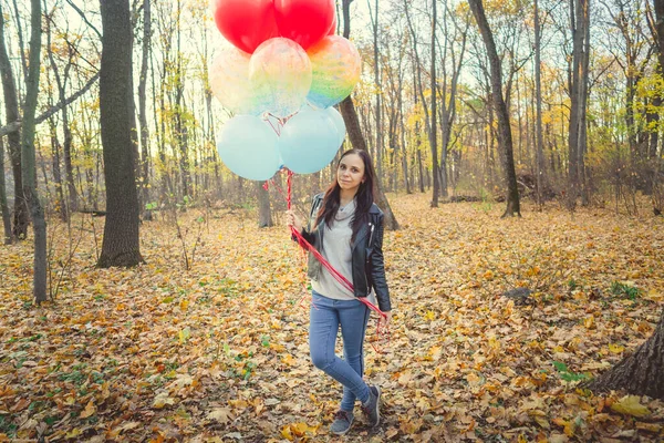 Una joven hermosa mujer en ropa casual está sosteniendo los globos de colores a pie en el bosque de otoño . — Foto de Stock