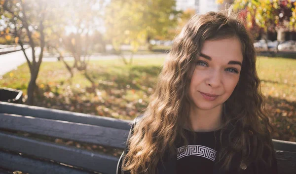 Uma jovem mulher bonita em roupas casuais está sentada em um banco no parque de outono . — Fotografia de Stock