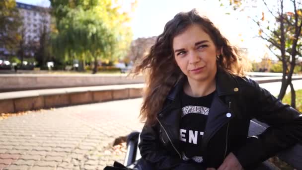 Uma jovem mulher bonita em roupas casuais está sentada em um banco no parque de outono . — Vídeo de Stock