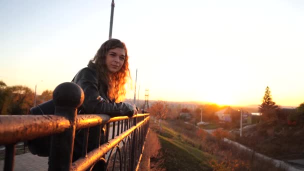 Молодая красивая женщина в кожаной куртке с черным рюкзаком стоит на мосту на фоне городского округа. — стоковое видео