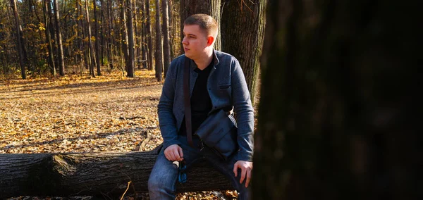 Homem sentado em madeiras de primavera. Masculino em sessão externa em log caído na floresta rural olhando para longe na luz do sol — Fotografia de Stock