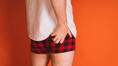 Male body in the plaid boxers and in white shirt on orange background. Mans hand scratches his ass.
