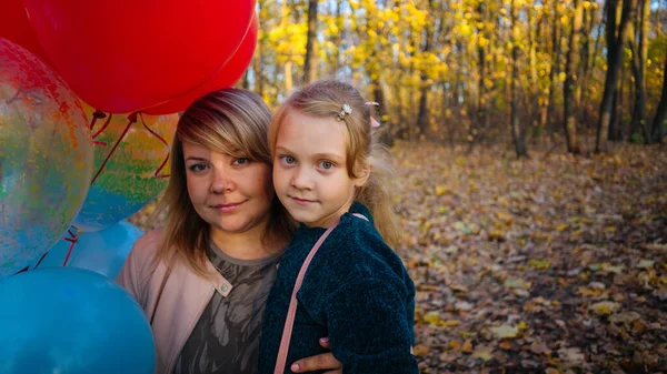 Eine schöne junge Mutter umarmt ihre kleine entzückende Tochter und hält die bunten Luftballons auf einem Wandspaziergang im herbstlichen Wald. — Stockfoto