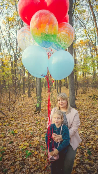Eine schöne junge Mutter umarmt ihre kleine entzückende Tochter und hält die bunten Luftballons auf einem Wandspaziergang im herbstlichen Wald. — Stockfoto