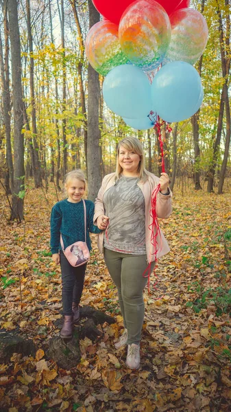 Mutter und kleine Tochter gehen händchenhaltend durch den herbstlichen Wald. Das Kind hält einen großen Strauß der bunten Luftballons in der Hand. — Stockfoto