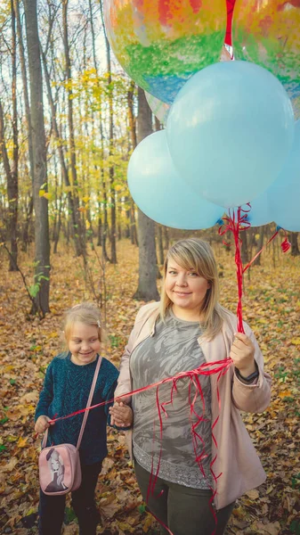 Mutter und kleine Tochter gehen händchenhaltend durch den herbstlichen Wald. Das Kind hält einen großen Strauß der bunten Luftballons in der Hand. — Stockfoto