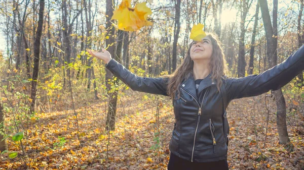 Eine schöne junge Frau in legerer Kleidung warf die gelben Blätter in den herbstlichen Wald. Das Laub fliegt in verschiedene Richtungen und die charmante Frau lacht fröhlich. — Stockfoto