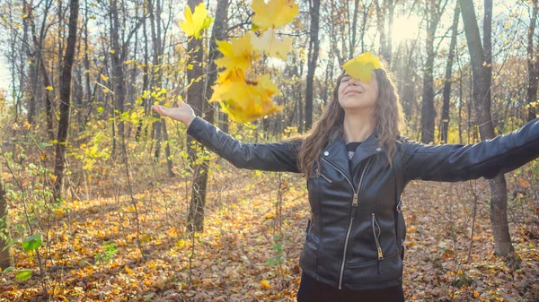 Eine schöne junge Frau in legerer Kleidung warf die gelben Blätter in den herbstlichen Wald. Das Laub fliegt in verschiedene Richtungen und die charmante Frau lacht fröhlich. — Stockfoto