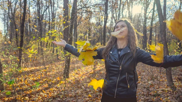 Eine schöne junge Frau in legerer Kleidung warf die gelben Blätter in den herbstlichen Wald. Das Laub fliegt in verschiedene Richtungen und die charmante Frau lacht fröhlich. — Stockfoto