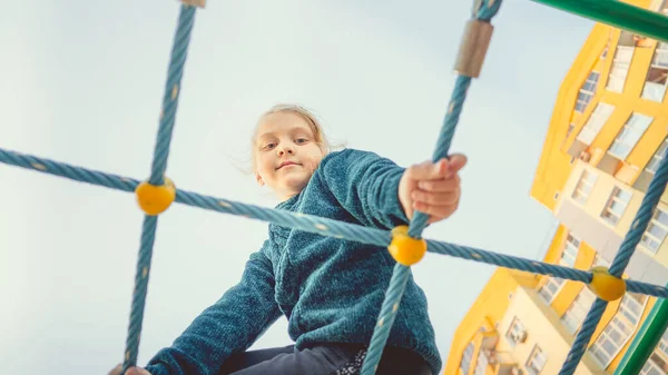 A charming little girl in casual clothes sits on the grid and looks at camera on playground in the urban district. — Stock Photo, Image