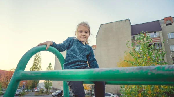 Ein charmantes kleines Mädchen in lässiger Kleidung sitzt auf einer Metallkonstruktion im Hof. — Stockfoto