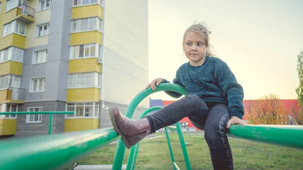 Ein charmantes kleines Mädchen in lässiger Kleidung sitzt auf einer Metallkonstruktion im Hof. — Stockfoto