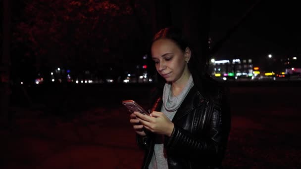 Een mooie vrouw typt een bericht op haar smartphone, tegen de achtergrond van een nachtstad. Vrouw die alleen staat 's nachts gebruikt haar telefoon om te communiceren op het internet. — Stockvideo