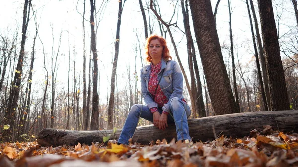 Mujer sentada en madera en el bosque de otoño. Mujer joven con pelo rojo vestida de civil sentada sobre troncos caídos en el bosque rural mirando hacia atrás bajo la luz del sol. — Foto de Stock