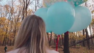 Genç bir anne ve küçük sevimli kızı renkli balonlarıyla sonbahar ormanlarında yürüyorlar..