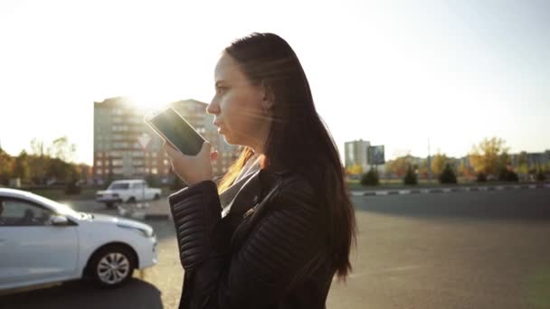 En ung kvinna står på bakgrunden av staden och spelar in ljudmeddelanden i ett socialt nätverk på en mobiltelefon. — Stockvideo