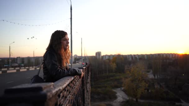 Una joven mujer guapa en una camisa de cuero con una mochila negra se levanta reflexivamente en el puente sobre el fondo del distrito urbano.. — Vídeo de stock