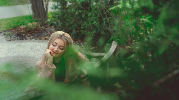 Un retrato de una hermosa niña, sentada en un banco cerca de la vegetación verde . —  Fotos de Stock