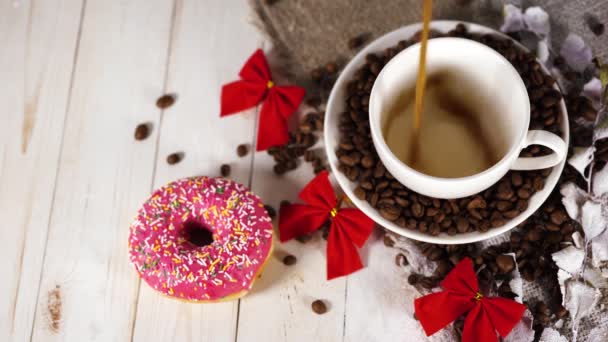 Ένα ζεστό φλιτζάνι καφέ με ένα ντόνατ και τους κόκκους καφέ στο τραπέζι. Σύνθεση του ένα νόστιμο καφέ και γλυκό σνακ. — Αρχείο Βίντεο