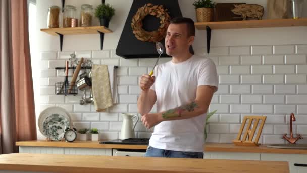 Um homem alegre canta com uma concha na cozinha. Um macho com uma boca aberta segura uma colher de servir em uma cozinha moderna e finge cantar — Vídeo de Stock