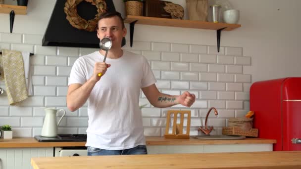 Un hombre alegre canta con un cucharón en la cocina. Un macho con la boca abierta sostiene una cuchara de servir en una cocina moderna y finge cantar — Vídeos de Stock