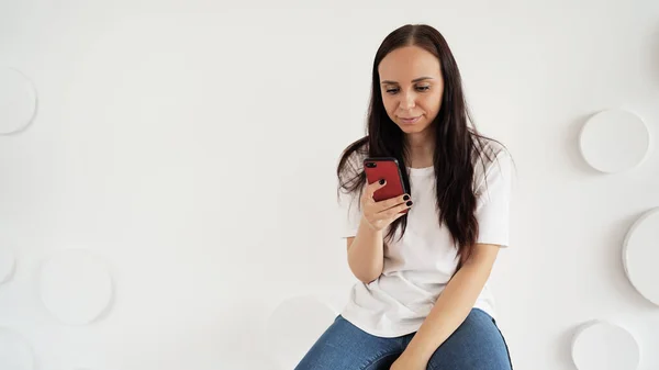 Young woman flipping chat in smartphone on background of white patterned wall. Adult female communicating on internet in mobile phone