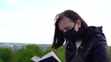 Siyah tıbbi maskeli ve günlük kıyafetleriyle tepede oturmuş bulutlu bir günde kitap okuyan genç bir kadın. Yetişkin dişi kırsalda tek başına dinleniyor..